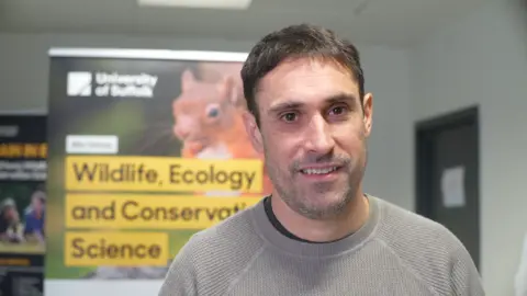 Jamie Niblock/BBC Dr Mark Bowler is standing in a lab. He has short brown hair and a greying moustache and beard. He is wearing a grey jumper.