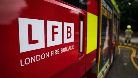 A red fire engine with LFB London Fire Brigade written in white lettering along the side of the vehicle. A firefighter is seen standing in the background, with a hose on the ground