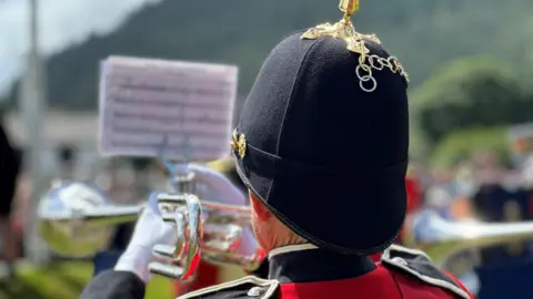 A trumpeter from the back. He is wearing a black helmet featuring a gold spike and white gloves. There is a music stand holding music attached to the trumpet.