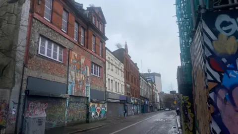 North Street in Belfast with empty shop units. Shutters are down with graffiti on them. The sky is grey.