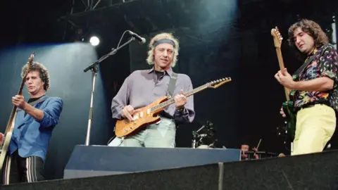Getty Images Dire Straits perform on stage. Three of the members are pictured each playing different guitars.