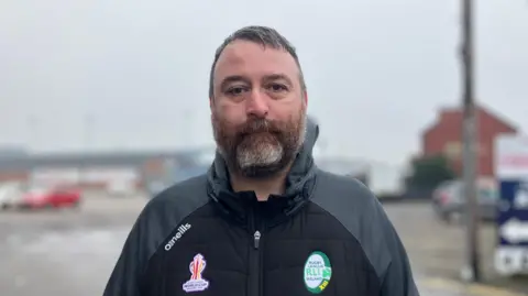 Elizabeth Baines/BBC A man with grey hair stands outside Featherstone Rovers ground. He wears a sports kit and has a grey beard