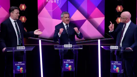 Three men in business suits stand at podium gesticulating at each other 