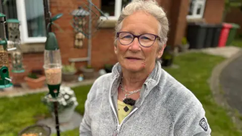 Avril Roddy in a garden. She is wearing a grey fleece with a yellow top. She has glasses and grey hair. There are bird feeders behind her.