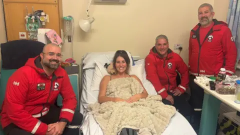 Women in hospital bed with mountain rescue standing around her 