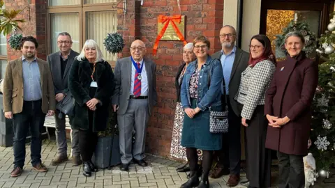 Swindon Borough Council A group of nine men and women stand either side of a gold plaque with a red bow on it. They are smiling for the camera and wearing smart clothing. The plaque is on a red-brick building and there are Christmas decorations dotted about behind the people.