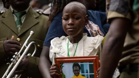 Badru Katumba/AFP Foto van de jongste dochter van Rebecca Cheptegei die huilt op haar begrafenis terwijl ze een foto van haar moeder vasthoudt in het Bukwo-district, Oeganda - zaterdag 14 september 2024.