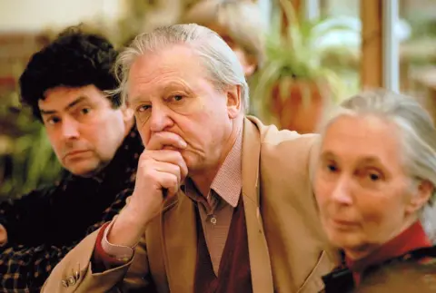 MonkeyWorld Jim Cronin, Sir David Attenborough and Jane Godall are sat together looking at the camera. Sir David, who is in full focus, holds a hand to his face. He is dressed in a smart jacket and shirt and has a serious expression on his face. 