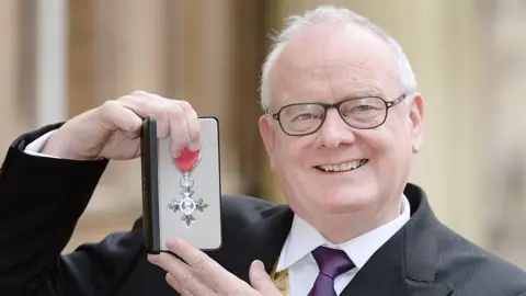 Dr Nigel O'Connor from Shrewsbury, a clean shaven man with grey hair and classes in a black suit and purple tie holds his Member of the Order of the British Empire, 