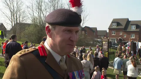 A man in a brown military uniform including a beret and with several medals is smiling as he is interviewed at the park, with many people attending the service in the background.