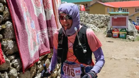 Shaunna Burke A woman dressed in purple with a pink t-shirt and black rucksack, wearing sunglasses and a running number. She is in what looks like a Nepalese mountain village. 
