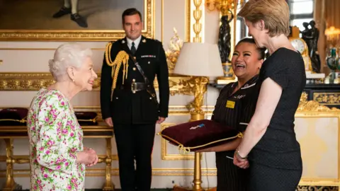 PA Media Queen Elizabeth II standing infront of May Parsons smiling, at Buckingham Palace alongside two staff members at the palace 