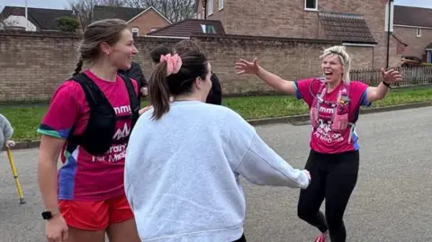Alice Draper A runner in a pink spreads her arms wide to greet two women during a marathon challenge. 