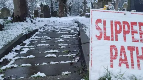 A Man For All Seasons/BBC Weather Watchers A snow-covered Slippery Path sign stands in front of a snowy graveyard