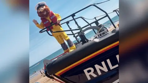 A person is wearing a bright yellow outfit with a red RNLI lifejacket and a mask on their head with a big brown beard and moustache and ship captain's hat. They are standing on an RNLI boat and waving to the camera. You can see a sunny beach and sea in the background.
