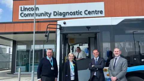 A wide angle shot of a bus outside Lincoln Community Diagnostic Centre. There are four people stood outside the bus. A driver is sat behind the steering wheel. 