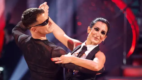 Dr Punam Krishan, wearing a black netted veil with a monochrome outfit with collar and tie, and Gorka Marquez, wearing a black long-sleeved top and black sunglasses, dance in hold on Strictly Come Dancing