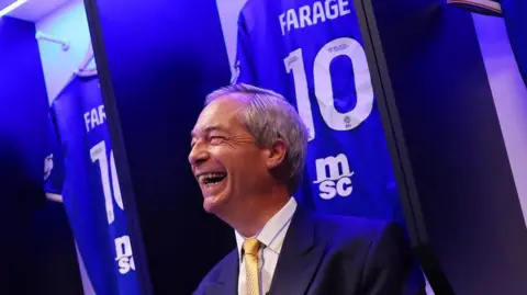 Nigel Farage in a navy suit and a yellow tie sits in Ipswich Town's home dressing room surrounded by personalised "FARAGE 10" Ipswich shirts. He is smiling and laughing as the picture is taken.