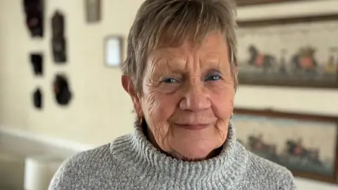 A woman in her 70s wearing a grey polo-neck jumper looks directly at the camera. She has short, light-brown hair and is standing in a room with pictures and artworks on a beige wall.