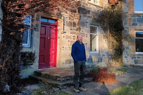 Assynt Foundation Lewis is standing outside a red door of  Glencanisp Lodge, a former Victorian hunting lodge.