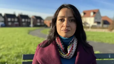 Bano is sat on a bench in a park. The sun is shining and she is wearing a maroon coat with a pattered scarf and blue jumper. She has shoulder length brown hair. Houses can be seen further back in the distance behind her.