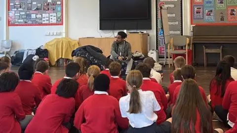 Breaking Barriers A group of primary‑school children in red jumpers sit on the floor of a bright school hall, all facing an adult who is speaking to them at the front. The room feels lively and colourful, with posters, displays and a large screen on the wall creating the sense of a busy learning space.