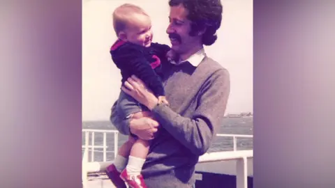 Family handout A toddler is smiling while being held by a man with short curly brown hair and a large moustache is smiling in this old family photo taken around the early 1980s. They are standing next to the sea.