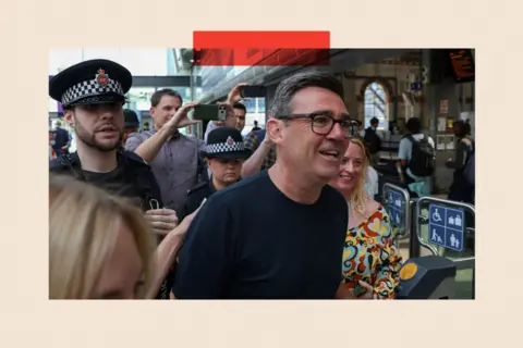 Reuters Newly elected Makerfield MP and former Greater Manchester Mayor Andy Burnham walks at Manchester Piccadilly station as he leaves for London, on 22 June 2026. He is a white man with dark greying hair and dark-framed glasses, and he wears a dark T-shirt. Behind him are two police officers. Two passers by are taking photographs of him with their phones.