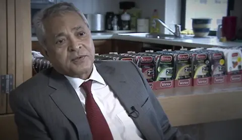 A man in a suit and red tie, sat down giving an interview