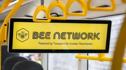 A yellow sign with the words 'Bee Network - powered by Transport for Greater Manchester' written in black. The sign is suspended from the roof of a bus, with seats, the bus windows and hanging straps seen in view. 