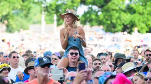 A music festival crowd is pictured head on. In the centre of the image a blonde woman wearing a blue dress is sat on a man's shoulders and towering over everybody else. 