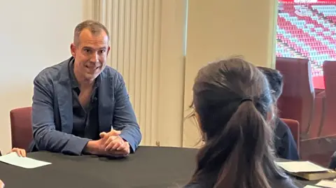 A middle-aged man wearing a blue jacket and black shirt, sitting at a table facing two children, who are sitting with their backs to the camera