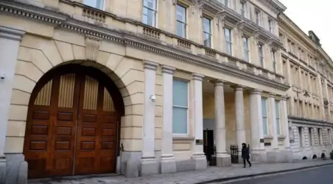 A grand white building with heavy set brown doors and pillars down the side of the building.