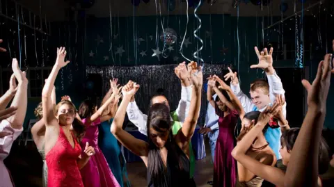Getty Images A stock photo of teenagers (15-18) in formalwear dancing at prom, arms raised. Streamer decorations hang from the ceiling.