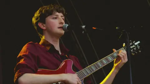 Ailis Cross-Gorman A man stands on stage singing into a microphone. He is playing a pink electric guitar and wearing a burgundy shirt.