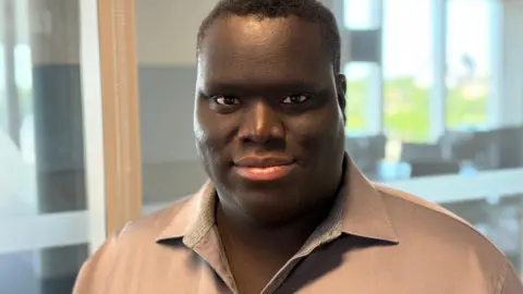 Kwaku Adje is looking directly at the camera in a brown shirt