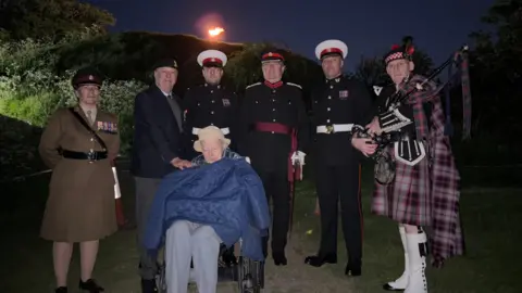 Cambridgeshire County Council Mr John Hayward is pictured in a wheelchair wearing a hat with a coat over his arms. behind him is a veteran, bagpipes player and other people dressed in military uniforms. Its dark and in the background the flame from the beacon can be seen on top of the hill. 