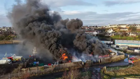 Fife Jammer Locations Thick black smoke and bright flames rise from a fire at Methil Harbour, engulfing heavy goods vehicles near a fenced industrial area. Fire engines and hoses are visible on the road, with houses and buildings in the background.
