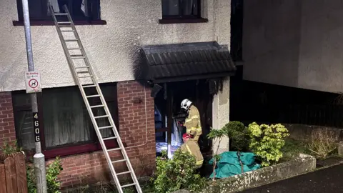 BBC A home showing signs of smoke damage with two fire fighters standing in uniform at the door. 