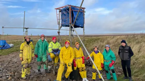 Eight people in caving suits are standing by a rig, which is a hoisting and counterweight tool called a Shadoof.