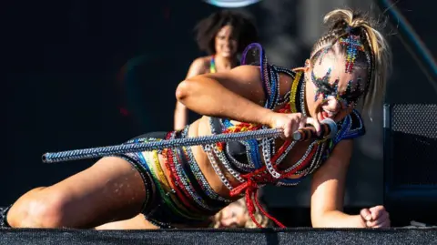 Getty Images JoJo Siwa lying connected  a signifier    singing and performing astatine  euphony  festival Mighty Hoopla successful  London successful  2024. She's wearing a achromatic  and brightly-coloured outfit covered successful  metallic  chains and holding a matching microphone. She has acheronian  oculus  make-up which is surrounded by multi-coloured jewels.