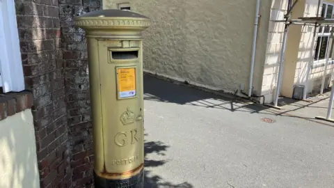 Gold post box next to a wall. A building is to the right of the image.