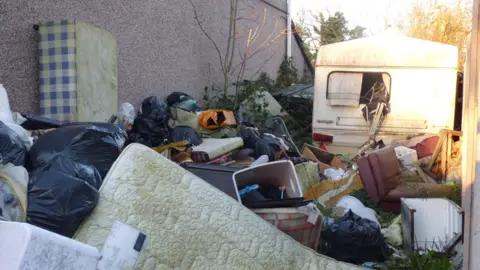 Piles of rubbish stacked up at the side of a property. The image shows an abandoned caravan to the back right of the picture. In the foreground are mattresses, bed bases, a sofa, black bin bags and various detritus. 