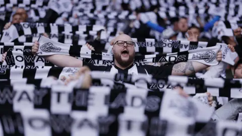 Reuters A sea of black and white scarves held aloft, with one man visible in the centre. He is bald, bearded, wearing glasses, and appears to be singing or chanting.