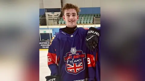 Rob Lamey Rufus Dawson training for the Great Britain Futures U16 team in summer 25. Rufus is wearing a blue and red jersey which has a lion on the front and a union jack flag underneath. He is wearing black hockey gloves and is standing on the ice.