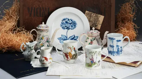 Woolley and Wallis A professional, advertisement-style photograph of the white place with a blue flowering auricula painted on it propped up against a wooden box with wood wool around it. In front of the plate are two tea pots and give cups or mugs. They are all white with various designs on them. Underneath them is various pieces of paperwork. 