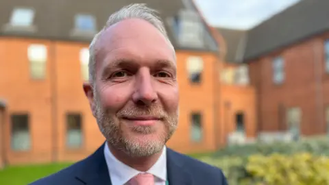 Jo Thewlis/BBC Steve James has short grey hair with a short beard and moustache.
He is wearing a blue suit with a white shirt and pink tie.
Behind him is the red bricks of The Link building and some green bushes.