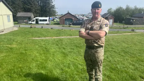 A man wearing an Army uniform is standing on grass with his arms crossed.