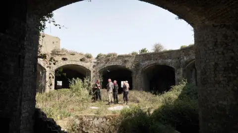 BBC View taken from inside an archway at a Victoria fort. It shows an open centre space with arches around the space. A handful of people are stood in the open space, which is filled with overgrown weeds