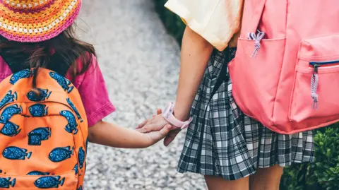 Getty Images Two young children walk hand in hand with back-packs on along a pathway towards a nursery